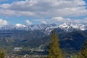 Snow-covered mountain range beneath soft blue sky; forested slopes and winding road add quiet...