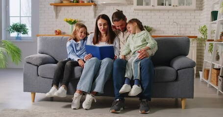 Happy loving family at home together, father, mother, kids reading book, parents with cute little child daughters, son smiling mum, dad telling small kids funny fairy tale story, sitting on sofa home - Powered by Adobe