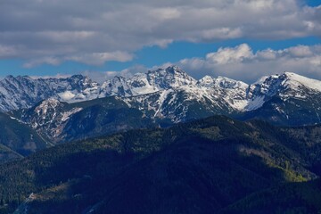 Snow-capped mountain range rises behind forested hills under partly cloudy skies; dramatic natural contrast shapes a striking scene.
