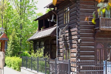 Rustic wooden house with log exterior and balcony sits behind metal fence, framed by lush trees and midday sunlight.