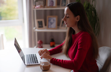 Young woman in a red sweater working on a laptop at home with a cup of coffee, focused on her work, natural daylight, cozy workspace.