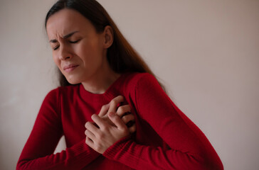 Young woman in a red sweater clutching her chest with eyes closed, expressing severe chest pain or heart attack symptoms, concept of heart health, stress, emergency.