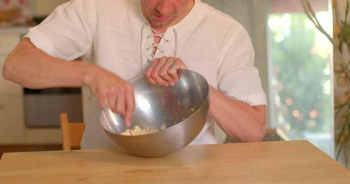 An individual is preparing delicious food by skillfully mixing various ingredients in a metal bowl using a spoon, all while positioned at a kitchen table filled with culinary tools