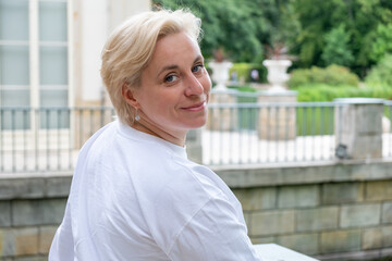 Midlife woman with short blonde hair smiles while sitting outdoors near a serene water feature,...