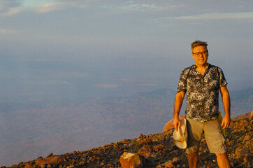A man stands atop a rocky hill, holding a hat, as he enjoys a stunning, expansive view during a calm sunset. The open, natural scenery symbolizes tranquility and adventure. 35mm film