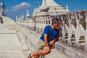 A Man in casual attire sitting on a historic building's rooftop, surrounded by architectural beauty and basking under a bright blue sky. Taken on 35mm film.