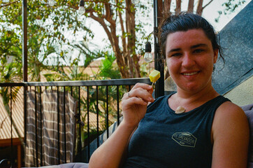 A cheerful woman seated outdoors enjoys a cubed pineapple snack on a patio surrounded by greenery,...