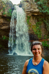 A cheerful woman wearing a blue shirt stands before a stunning waterfall surrounded by lush greenery, portraying joy and adventure in an outdoor natural environment. Taken on 35mm Film.