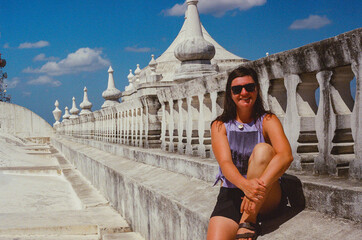 Woman seated at the top of an intricate white architectural structure under a bright sunny sky, radiating relaxation and enjoyment during clear weather. Taken on 35 mm Film.