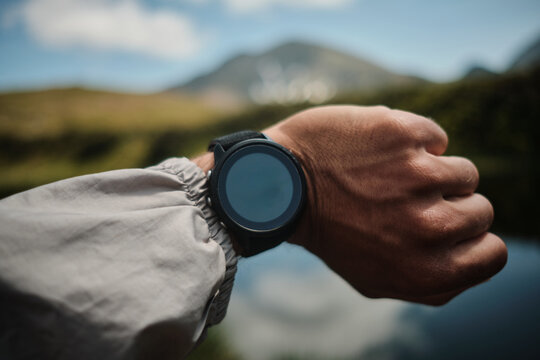 Close-up of a person wearing a smartwatch outdoors, with a blurred mountain landscape in the background. The image evokes a sense of adventure and technology.