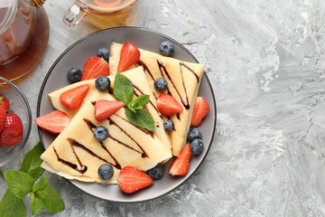 Tasty crepes with berries, chocolate sauce, mint and tea on gray textured table, flat lay. Space for text