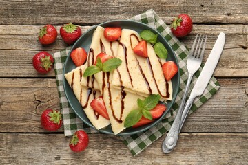 Tasty crepes with strawberries, chocolate sauce and mint on wooden table, flat lay