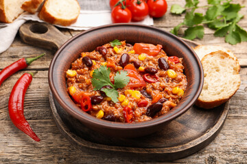 Tasty chili con carne in bowl, ingredients and bread on wooden table, closeup
