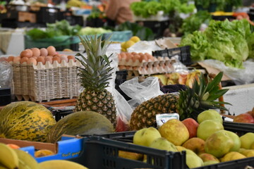 Fresh pineapples, melons, and apples on display at a local market stand, with leafy greens and other produce in the background.