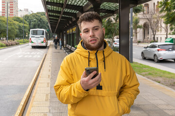 young Latino man waits for the bus at the stop with his smartphone in his hand. travel fare increase