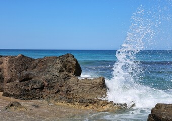 Wave explodes against rocky Crete coast with sea spray and turquoise water under blue sky. Dynamic...