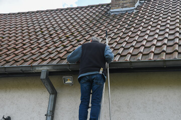 Professional roofer cleaning residential gutter while balancing on metal ladder, wearing work attire and checking roof condition under bright sunlight