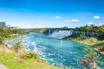 A scenic view of Niagara Falls from the Canadian side, showing the American Falls, Rainbow Bridge, and tour boats navigating the turquoise river below on a bright summer day.