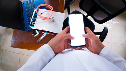 Personal view perspective of  doctor holds a smartphone with white screen at their desk, surrounded by medical tools, a laptop, and office supplies.