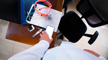 Personal view perspective of  doctor taking smartphone with white screen as copy space from their desk, surrounded by medical tools.