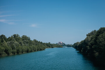The photo features a calm river flowing through lush green banks under a clear blue sky. In the distance, Ptuj Castle sits atop a hill, providing a picturesque focal point framed by trees and water.