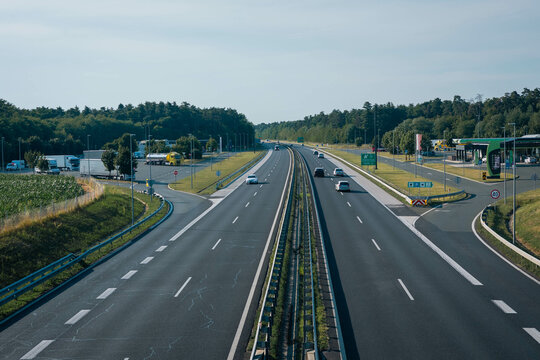clean, modern highway with light traffic, surrounded by green forests and fields. There s a service area with a gas station, trucks, and cars, under a clear blue sky.