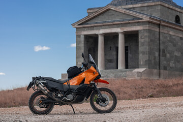orange adventure motorcycle stands poised on a gravel path along Majstorski Put, its rugged design contrasting beautifully with a historic stone building featuring elegant columns. Under a vast, clear