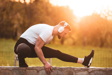 A young fit man listening to music on wireless headphones while doing stretching exercises before running or workout in the park at sunset. Outdoor fitness preparation, active lifestyle concept.