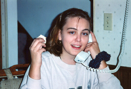 Circa 1996 - teenage girl talks on a landline phone with her friend while eating. Scrunchie on wrist, typical 90s highschooler.