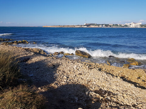 A rocky shoreline with waves crashing against the stones. The clear blue sea meets the sky, creating a serene coastal landscape.