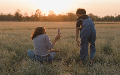Mother holding smartphone captures photo of her young son during a peaceful sunset in a flower field. Boy is standing while mother sits on the grass holding a smartphone, in the golden hour light.