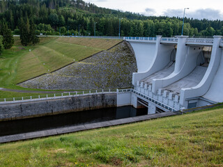 Hydroelectric dam with curved spillways, grassy hillside, and water outlet channel. Forest in...