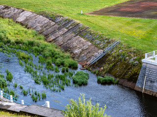 River flows below a dam along a concrete slope with metal stairs. Green vegetation grows in water....