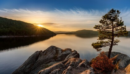a lone tree stands on a rocky cliff overlooking a calm lake at sunset surrounded by forested hills
