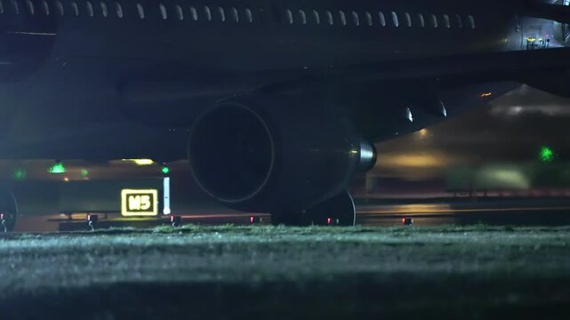 Close view of airplane engine and wing over taxiway sign M5 at night with lights and reflections on surface