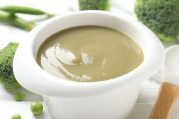 Baby food. Tasty puree in bowl, spoon, peas and broccoli on white wooden table, closeup