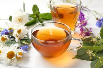 Aromatic herbal tea in glass cups, different flowers and green leaves on white wooden table, closeup