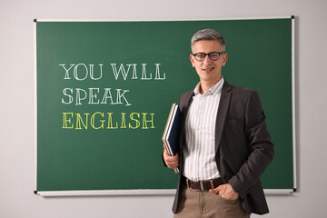 Teacher near green chalkboard with text You Will Speak English. Language course