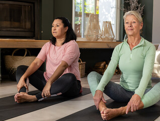 Diverse senior female friends practicing butterfly yoga in living room on yoga mats near fireplace