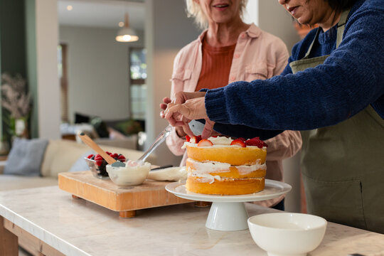 Diverse senior female friends decorating layered cake on kitchen island with cream and strawberries - Powered by Adobe