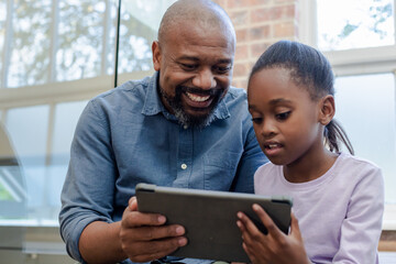 African American father and daughter sitting on couch by window at home sharing tablet content