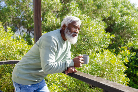 Senior African American man leaning on deck railing holding grey mug wearing sweatshirt, copy space