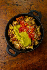 A cast iron skillet of loaded potatoes with shredded beef, melted cheese, guacamole, and pico de gallo; a hearty pub food appetizer perfect for sharing, served on a wooden table.