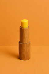 A mockup of a yellow lip balm stick in a wooden tube standing upright on a vibrant orange background for natural beauty