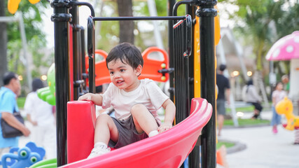A young Asian boy playing happily on a colorful playground slide in a park during daylight. He shows joy, curiosity, and energy while exploring the play area. 