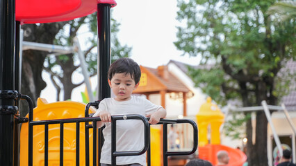 A young Asian boy playing happily on a colorful playground slide in a park during daylight. He shows joy, curiosity, and energy while exploring the play area. 
