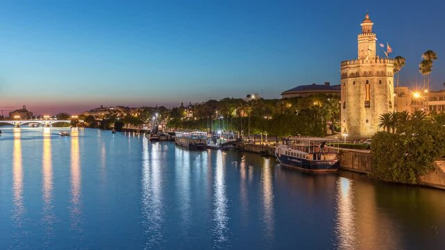 Tour boat floating along the Guadalquivir River near Torre del Oro watchtower in Seville, Spain. Day to night transition timelapse with city lights reflecting on water and scenic view of a waterfront