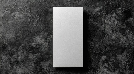 A vertically-oriented, blank white card rests on a dark, textured surface.  The card has a subtle, light texture visible. The background is mottled dark gray and black