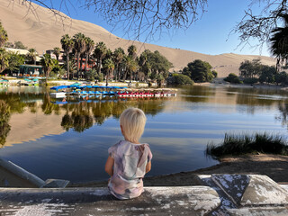 Little child or toddler sitting back to the camera and watching Huacachina lagoon surrounded by sand dunes desert, all reflected in the lake, Atacama desert, Ica, Peru, South America. 