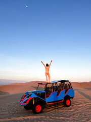 Woman standing on the roof top of buggy, that drives tourists around in desert area, while holding arm raised and watching beautiful arid landscape scenery Huacachina, Ica, Peru. Travel concept.